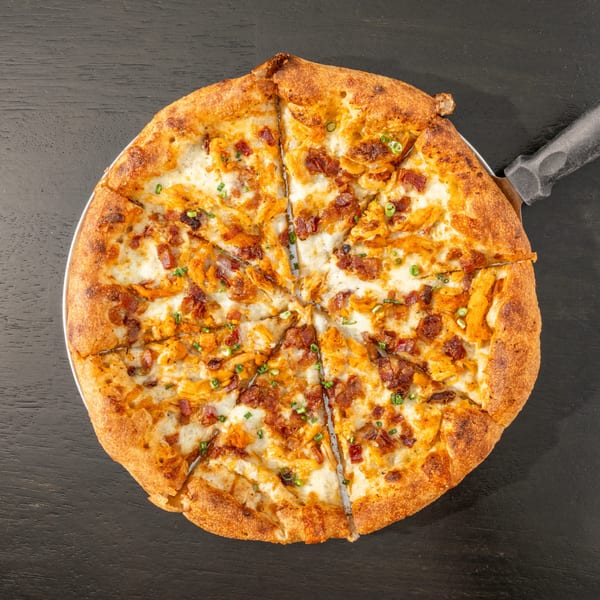 Overhead view of a sliced Black Hawk Brewery pizza with chicken, bacon, and chives on a dark table.