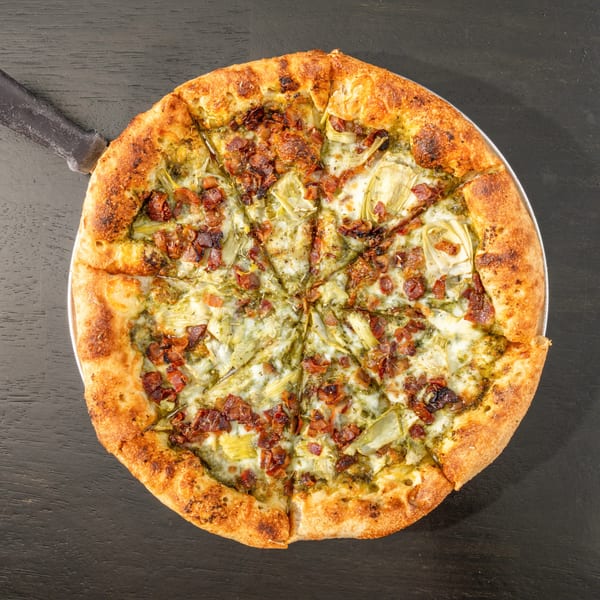 A Black Hawk Brewery pizza with pesto, artichokes, and bacon, sliced on a dark table.
