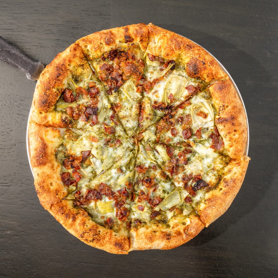 Overhead view of a Black Hawk Brewery pizza with pesto, artichokes, bacon, and cheese.