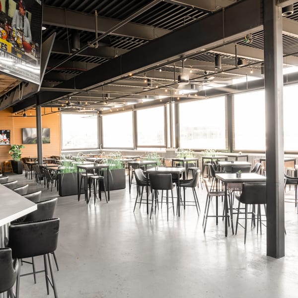 Spacious sports bar interior with high-top tables, bar seating, large windows, and a TV showing a game.