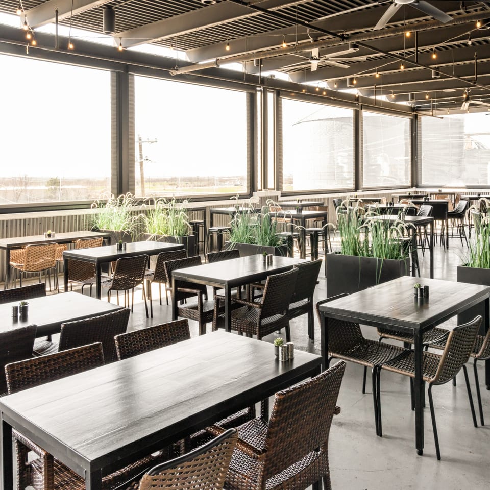 Black Hawk Brewery's spacious outdoor patio with dark tables, wicker chairs, and tall green plants.