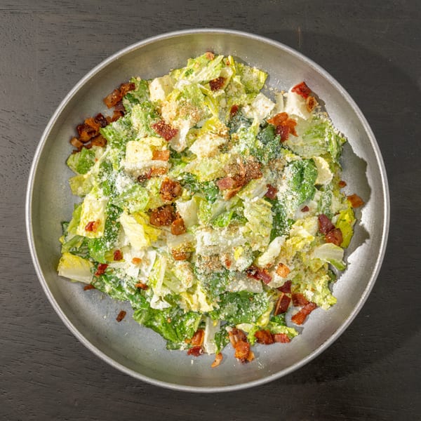 Overhead view of a Caesar salad with crispy bacon bits and grated cheese in a metal bowl.